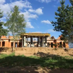 Surowy stan budynku z czerwonej cegły, w trakcie wznoszenia, z widocznym szalunkiem na stropie, wspartym na metalowych podporach, na tle błękitnego nieba z chmurami, otoczony drzewami iglastymi...