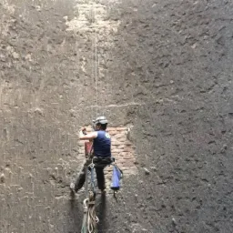 Pracownik w uprzęży, zabezpieczony linami, wykonuje prace na wysokości na szarej, chropowatej ścianie budynku z widocznym fragmentem muru z czerwonej cegły.