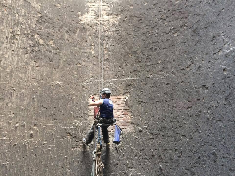 Pracownik w uprzęży, zabezpieczony linami, wykonuje prace na wysokości na szarej, chropowatej ścianie budynku z widocznym fragmentem muru z czerwonej cegły.