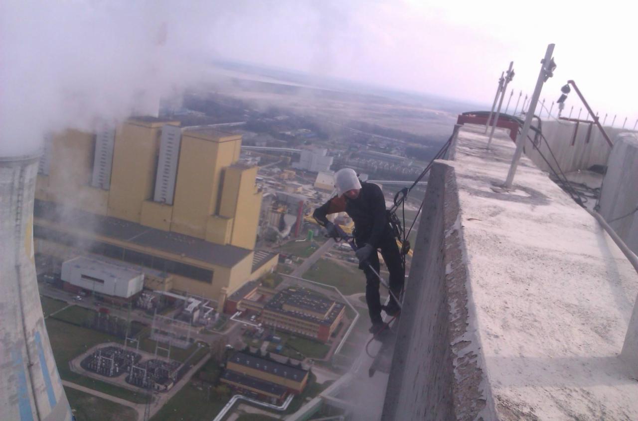 Pracownik wysokościowy na krawędzi dachu elektrowni, zabezpieczony linami, wykonuje prace konserwacyjne na tle rozległego krajobrazu miejskiego i wiejskiego.