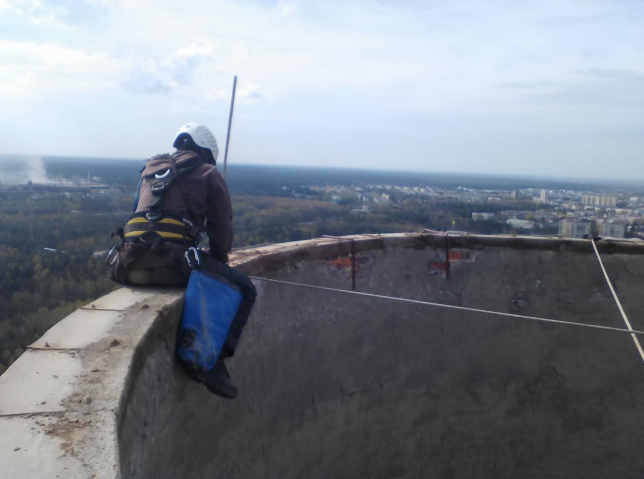 Pracownik w uprzęży i kasku siedzi na krawędzi betonowej konstrukcji na dużej wysokości, z widokiem na miasto i las w tle.