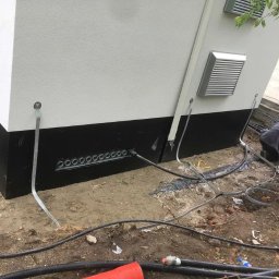 Spark System - Przyłącze kablowe do budynku z licznymi otworami wentylacyjnymi w fundamencie. Widoczne kable i uziemienie, instalator w trakcie pracy.