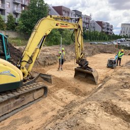 Spark System - Żółta koparka Ammann Yanmar w wykopie, pracownicy z niwelatorem i ubijarką, przygotowują teren pod budowę na tle budynków mieszkalnych.