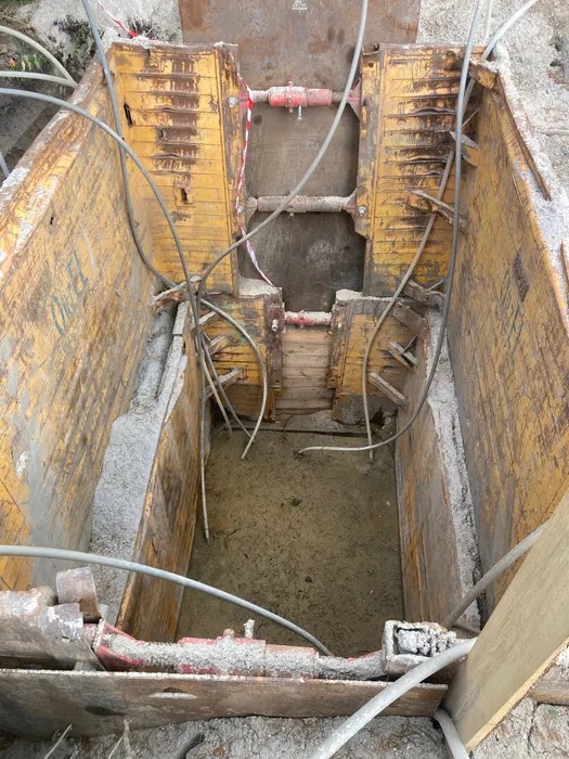 Głęboki wykop zabezpieczony żółtymi szalunkami stalowymi, widoczne rury i elementy konstrukcyjne. Dno wykopu wypełnione wodą z piaskiem.