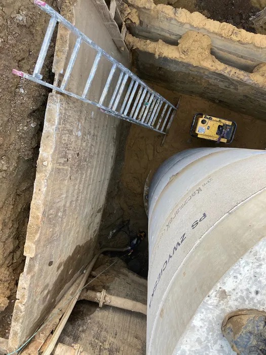 Głęboki wykop z betonowymi ścianami, wewnątrz rura kanalizacyjna, drabina i agregat prądotwórczy. Widoczny pracownik w dole wykopu. Ujęcie z góry.