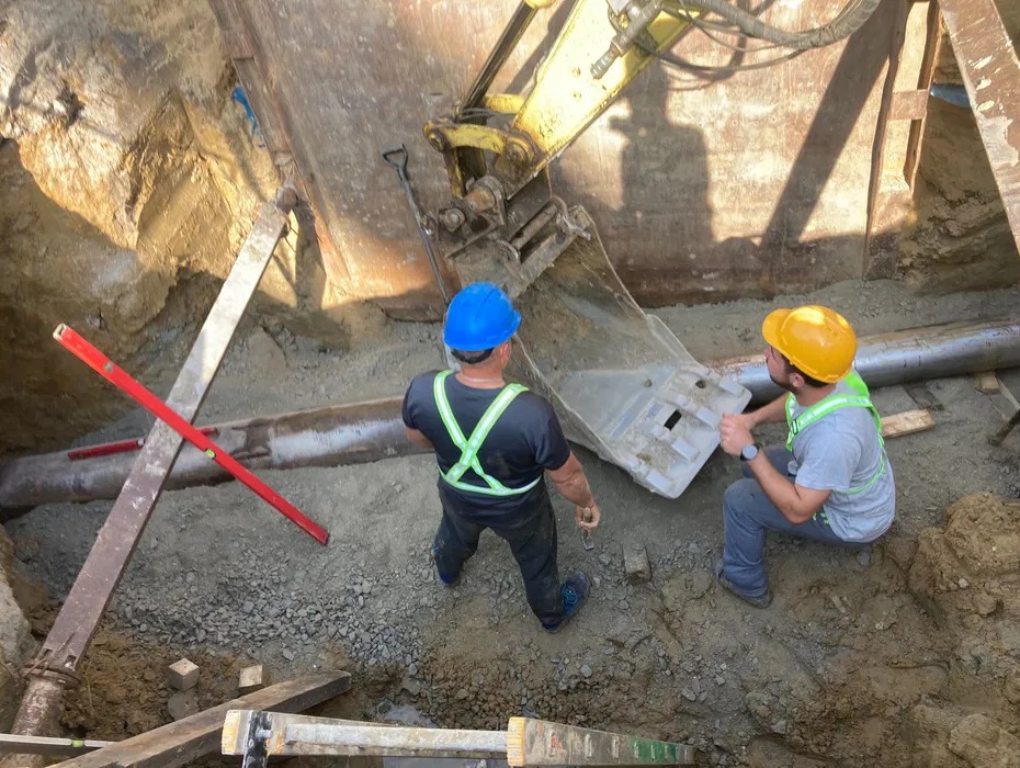 Dwóch pracowników w kaskach przy wykopie z rurą, obok koparka. Jeden z nich trzyma element konstrukcyjny. Widok z góry na plac budowy.