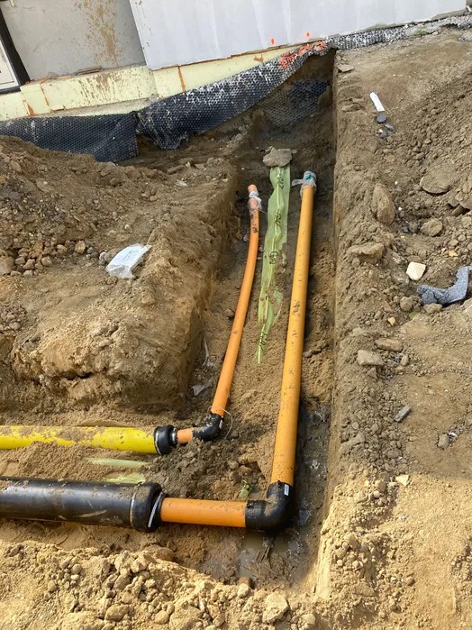 Wykop z ułożonymi rurami kanalizacyjnymi w ziemi, widoczne złączki i fragment budynku w tle. Rury w kolorach: żółtym, pomarańczowym i czarnym.