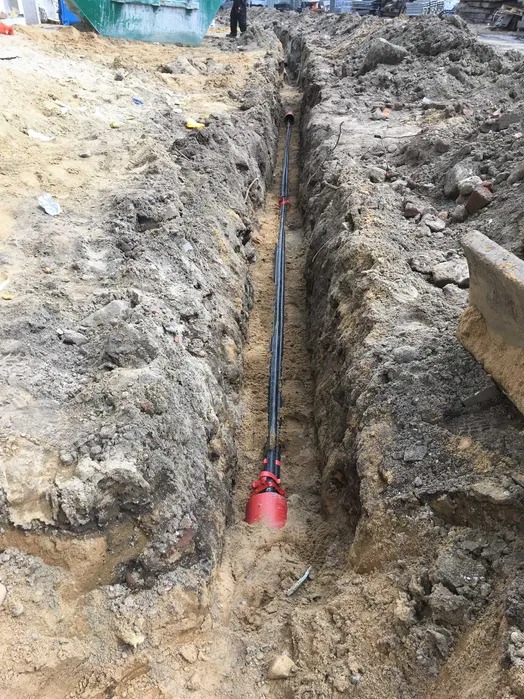 Wykopany wąski rów z ułożonymi rurami w czerwonych osłonach. Widok z góry na wykop w ziemi. Prace ziemne, instalacja podziemna.