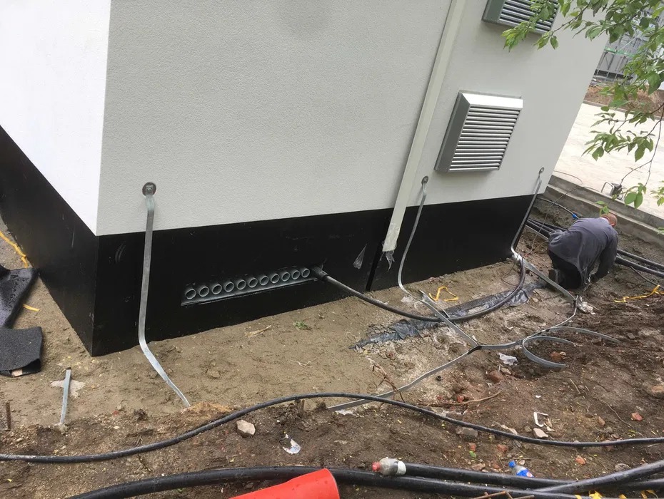 Przyłącze kablowe do budynku z licznymi otworami wentylacyjnymi w fundamencie. Widoczne kable i uziemienie, instalator w trakcie pracy.