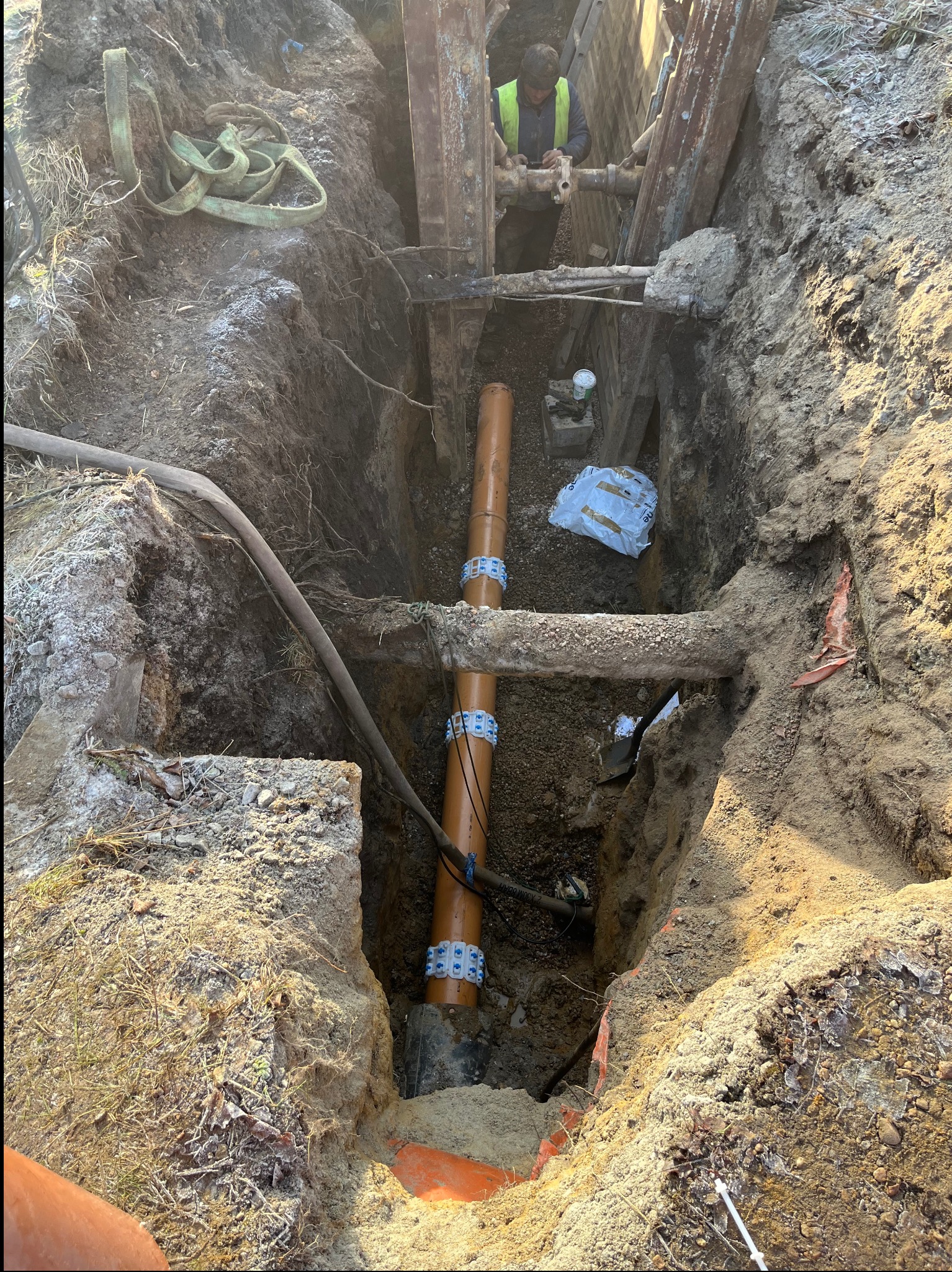 Głęboki wykop z pomarańczowymi rurami kanalizacyjnymi połączonymi niebieskimi złączkami. W tle pracownik w kamizelce odblaskowej przyłącza rury do instalacji.