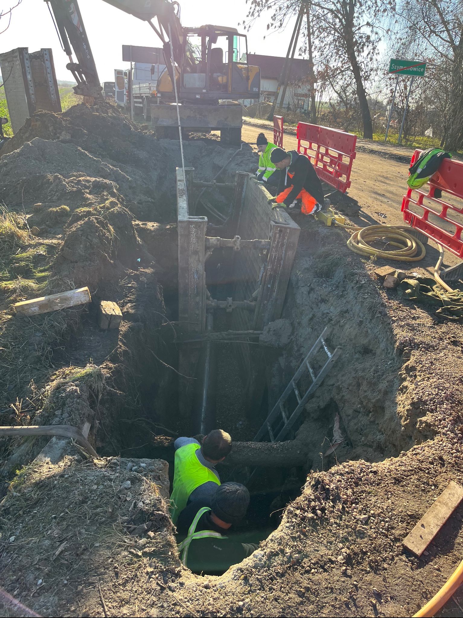 Głęboki wykop zabezpieczony stalową szalunką, ekipa w kamizelkach odblaskowych pracuje wewnątrz. Na górze koparka i tablica z nazwą miejscowości Szymanowice.