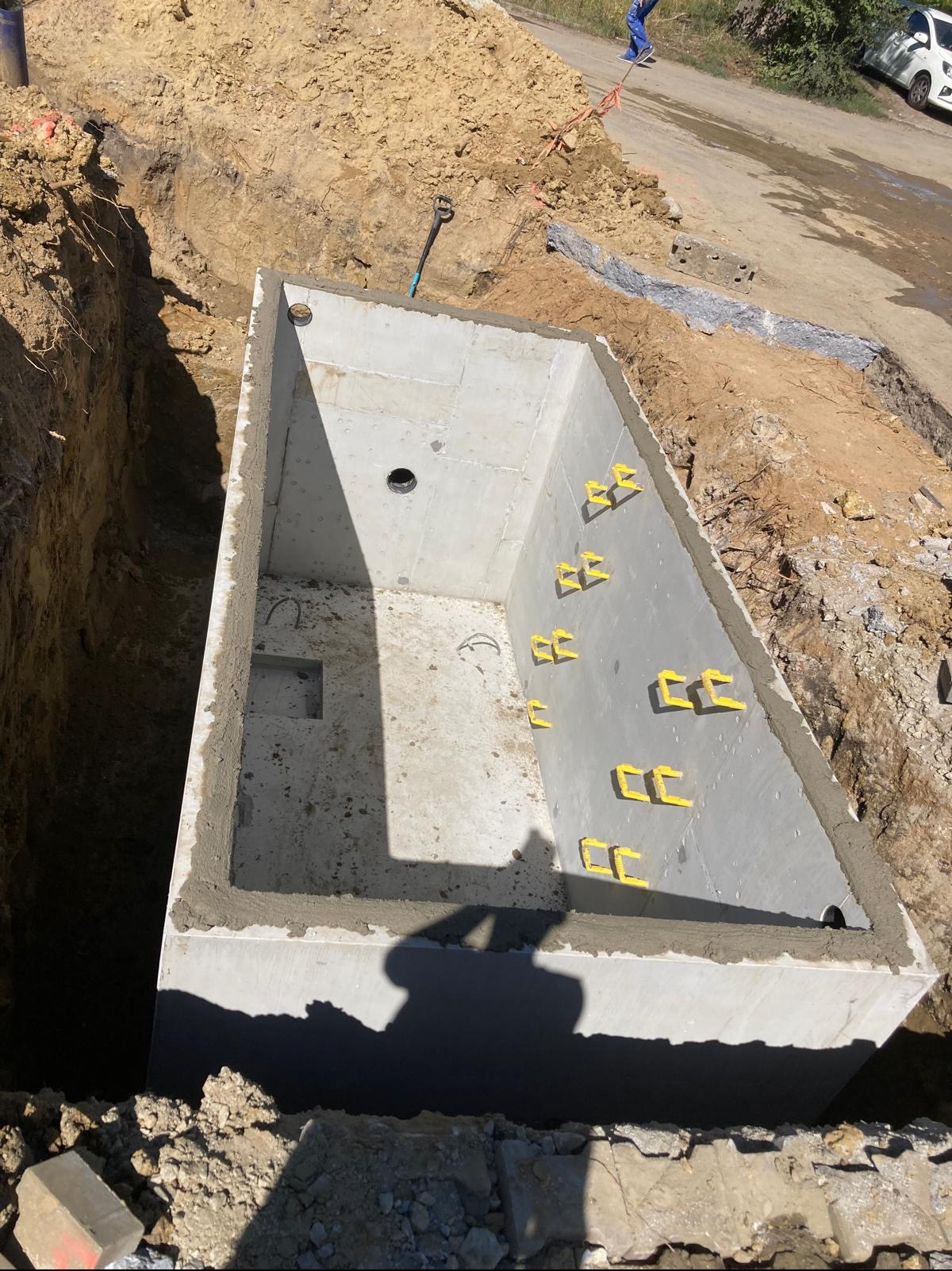 Betonowy zbiornik w wykopie, z żółtymi uchwytami montażowymi, widok z góry. Etap budowy szamba betonowego z wykończonymi krawędziami. W tle ziemia i fragment drogi.