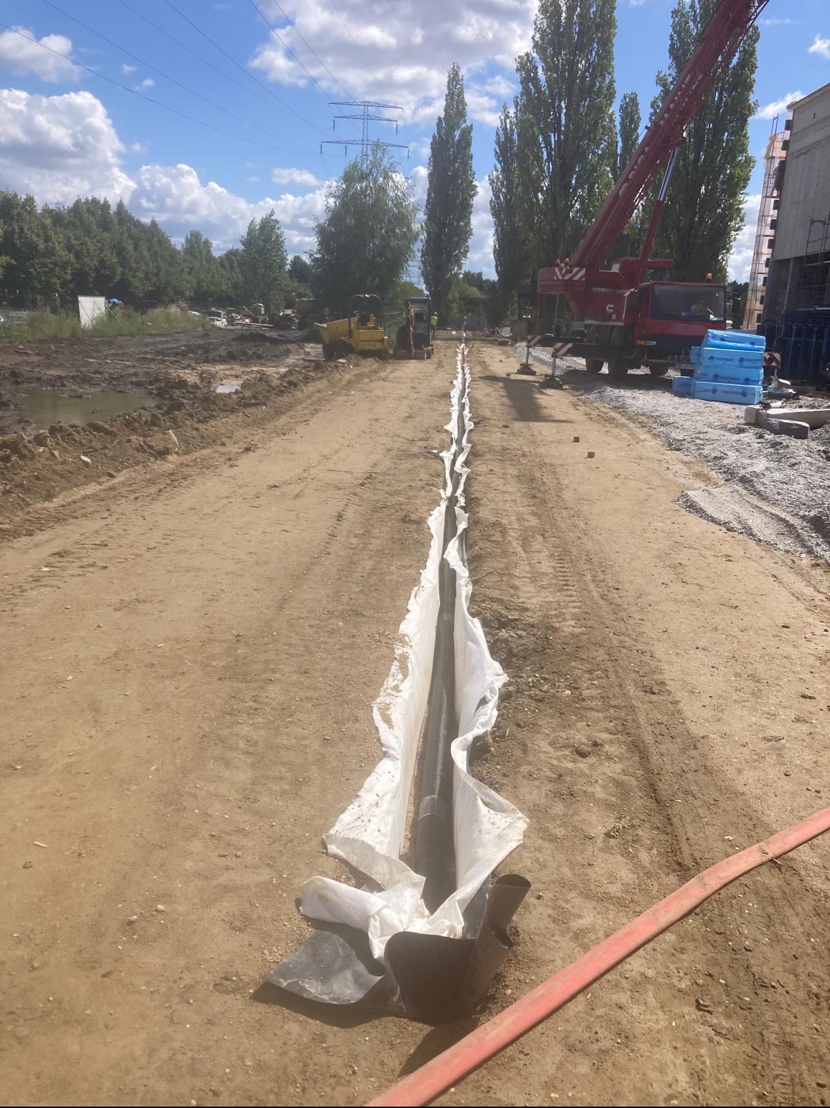 Długi wykop z rurą drenażową w białej geowłókninie, na tle maszyny budowlanej i żurawia. Widok z góry na plac budowy w słoneczny dzień.