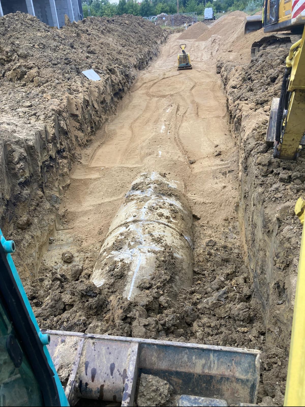 Wykop z ułożonym, dużym, betonowym elementem rurowym. W tle ubijarka i hałda ziemi. Widok z kabiny koparki na wykop.