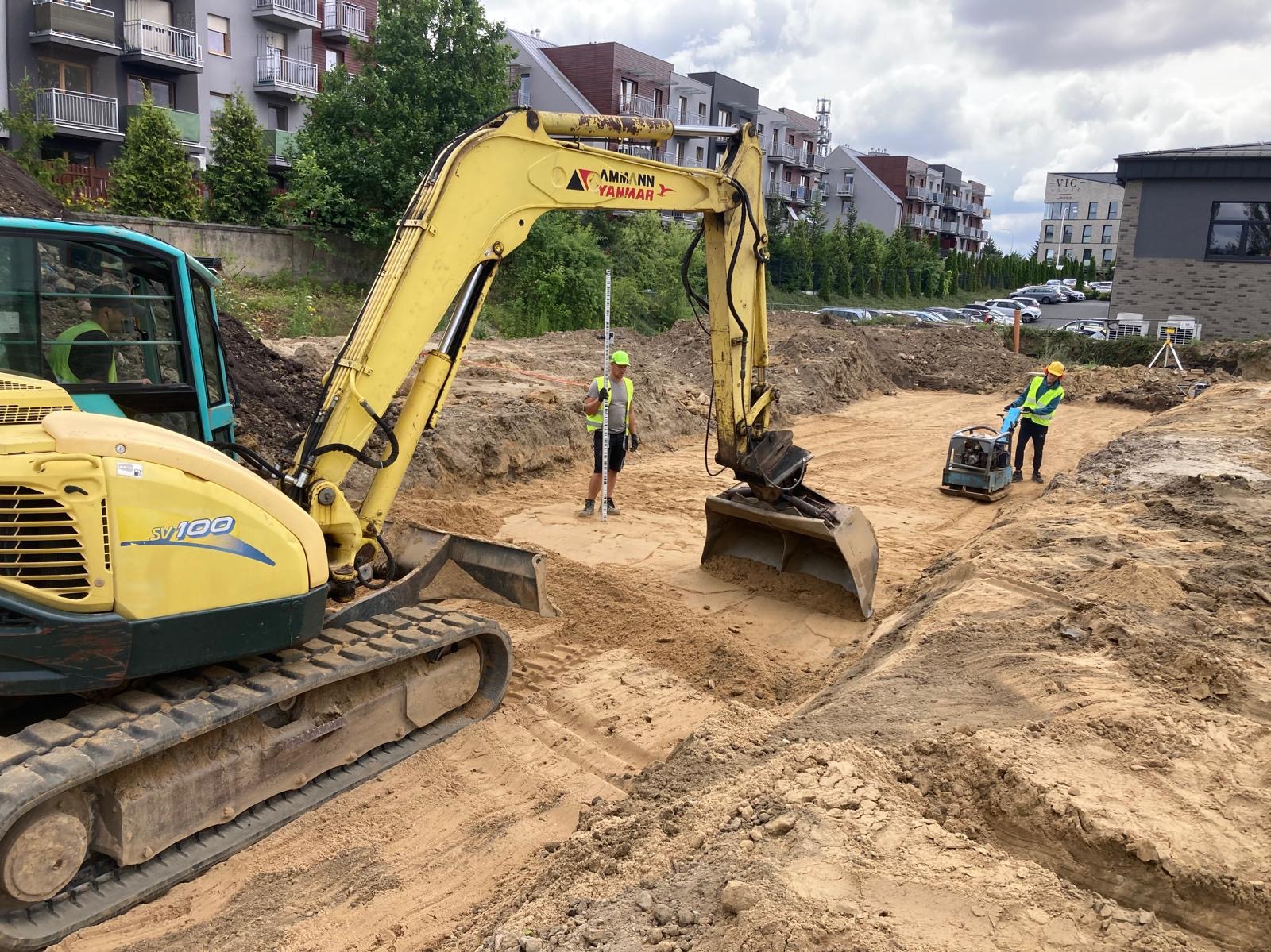 Żółta koparka Ammann Yanmar w wykopie, pracownicy z niwelatorem i ubijarką, przygotowują teren pod budowę na tle budynków mieszkalnych.