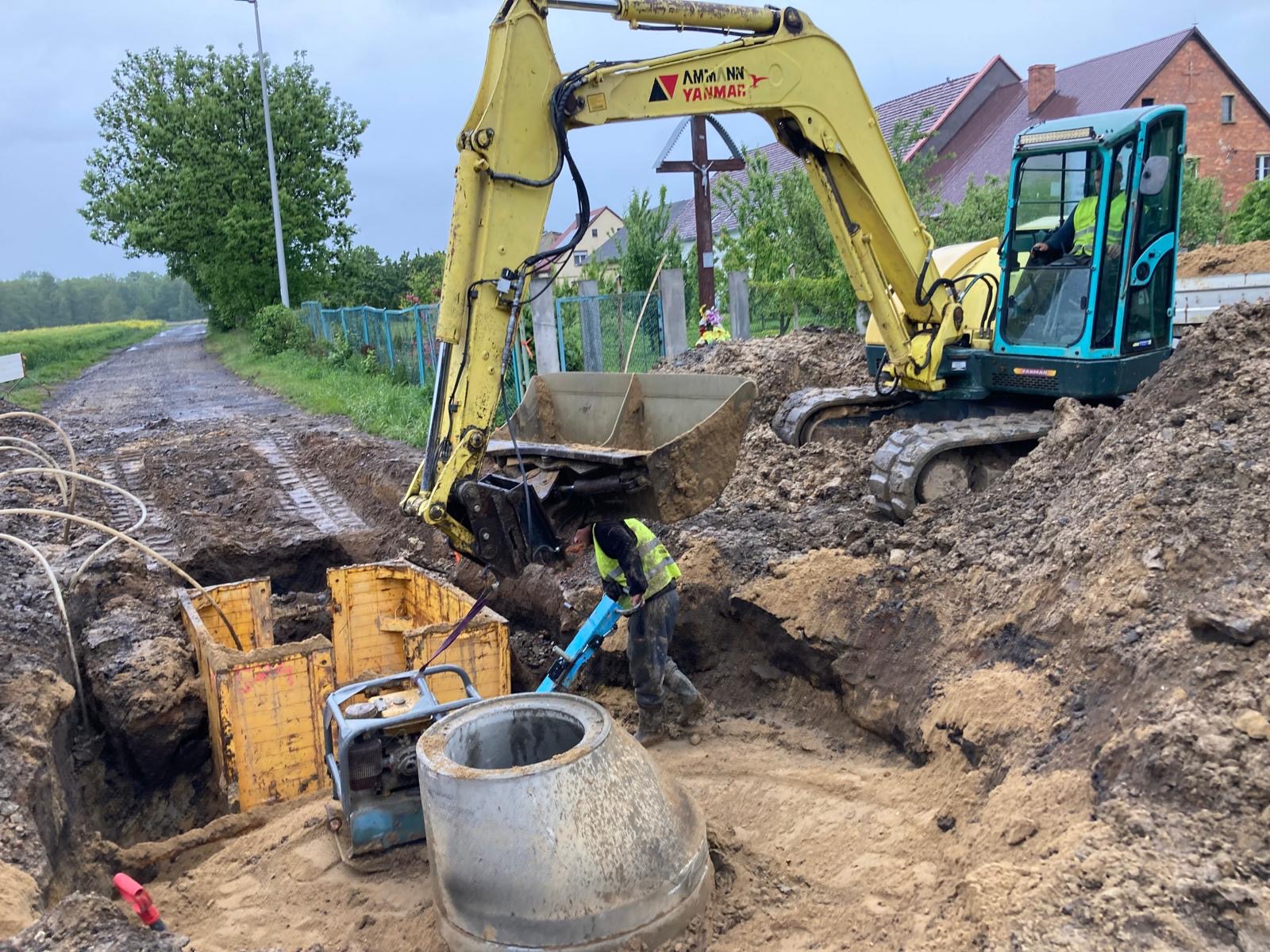 Żółta koparka Yanmar w trakcie prac ziemnych przy wykopie z żółtymi szalunkami. Pracownik używa młota pneumatycznego. W tle droga i zieleń.