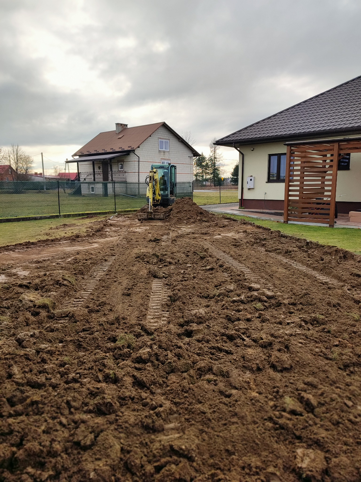 Żółta mini koparka na rozkopanym terenie działki, widoczne ślady gąsienic, w tle dwa domy jednorodzinne i pochmurne niebo.