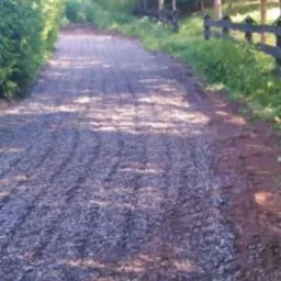 Długa, gruntowa droga wysypana drobnym kamieniem, otoczona zielenią i drewnianym płotem, w słoneczny dzień.