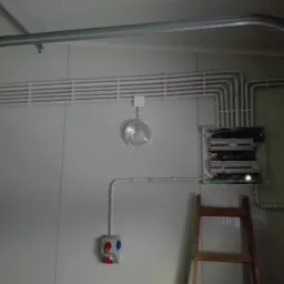 Rozdzielnica elektryczna z poprowadzonymi przewodami w białych rurkach, na ścianie z widoczną skrzynką z gniazdami i wyłącznikami, obok drabina.