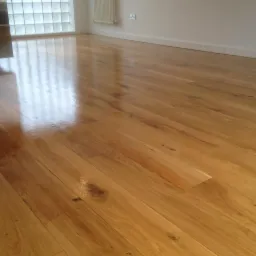 Błyszcząca, świeżo polakierowana podłoga z jasnego drewna w pustym pokoju z grzejnikiem i oknem z luksferami.