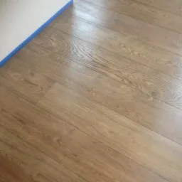 Ułożone jasnobrązowe deski podłogowe z widocznym rysunkiem słojów, zabezpieczone niebieską taśmą malarską przy ścianie.