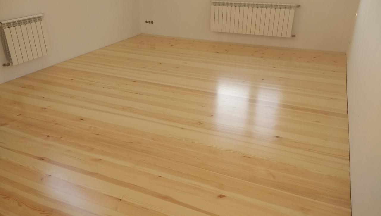 Jasne, świeżo ułożone deski podłogowe z widocznym usłojeniem drewna, w pustym pokoju z białymi ścianami i grzejnikami.