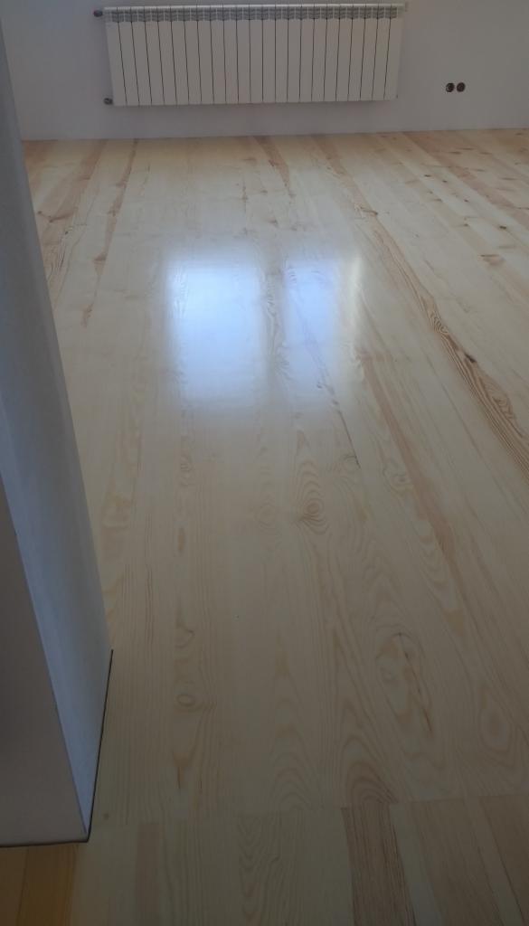 Jasne, lakierowane deski podłogowe z widocznym rysunkiem drewna, ułożone wzdłuż pomieszczenia, z białym grzejnikiem żeberkowym na ścianie w tle.