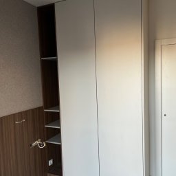 Meble Partyka Szymon Partyka - Wnętrze z dużą, białą szafą z przesuwnymi drzwiami, obok otwarta szafka z półkami i instalacją elektryczną w ścianie.