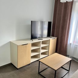 Meble Partyka Szymon Partyka - Nowoczesny salon z beżowo-drewnianą szafką RTV z otwartymi półkami, czarnym telewizorem, kawowym stolikiem z czarną ramą i brązowymi zasłonami w tle.