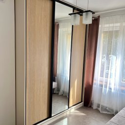 Meble Partyka Szymon Partyka - Trzydrzwiowa szafa wnękowa z przesuwnymi drzwiami: dwoje drzwi z okleiną drewnopodobną i jedne drzwi z lustrem, czarne obramowanie, jasne wnętrze pokoju z zasłonami i częściowo widocznym...