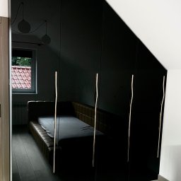 Meble Partyka Szymon Partyka - Czarna, błyszcząca szafa wnękowa z podłużnymi, złotymi uchwytami, odbijająca fragment sypialni z łóżkiem i oknem.