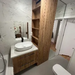 Nowoczesna łazienka z umywalką nablatową, drewnianą szafką z szufladami i wysoką szafką regałową z otwartymi półkami, lustrzane ściany, marmurowe płytki na ścianach.