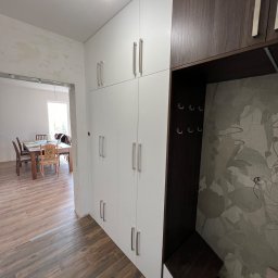 Meble Partyka Szymon Partyka - Biało-brązowa zabudowa meblowa z szafkami i otwartą wnęką z wieszakami, widoczna w przedpokoju z drewnopodobną podłogą i przejściem do jadalni w tle.