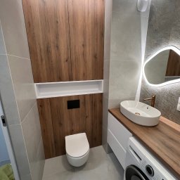 Meble Partyka Szymon Partyka - Nowoczesna łazienka z drewnianymi akcentami: szafka wisząca nad toaletą, umywalka nablatowa na blacie z efektem drewna, oświetlone lustro o nieregularnym kształcie i pralka pod blatem.