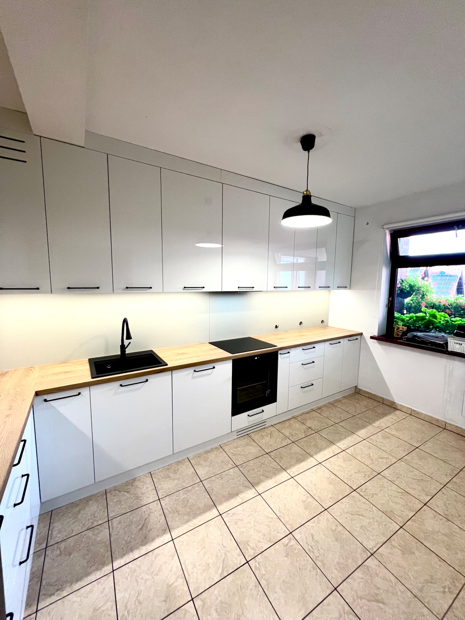 Nowoczesna, jasna kuchnia z białymi szafkami, drewnianym blatem i czarnymi akcentami: zlew, bateria, uchwyty, płyta indukcyjna i lampa wisząca.