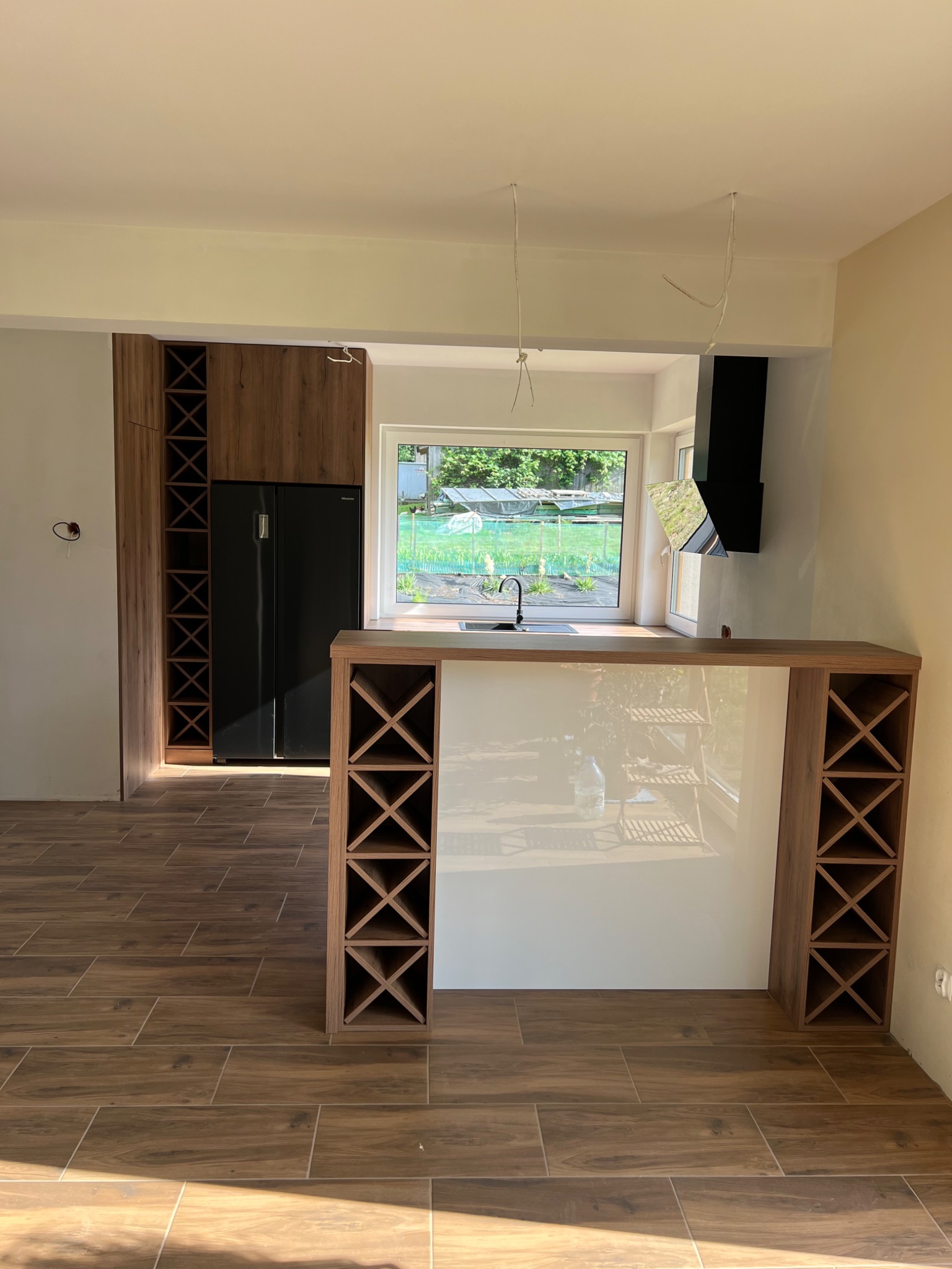 Nowoczesna kuchnia w trakcie wykańczania: drewniane szafki z miejscem na wino, czarna lodówka side-by-side, wyspa z białym frontem i drewnianymi regałami na wino, brązowe płytki imitujące drewno...