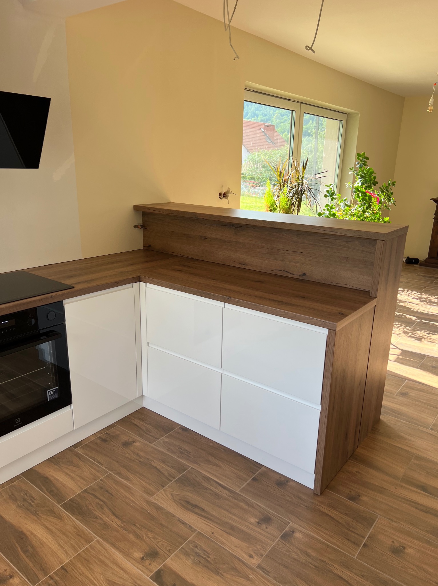 Nowoczesna kuchnia z białymi frontami szafek i blatem imitującym drewno, z widocznym piekarnikiem i płytą grzewczą, z oknem w tle i zielenią za nim.