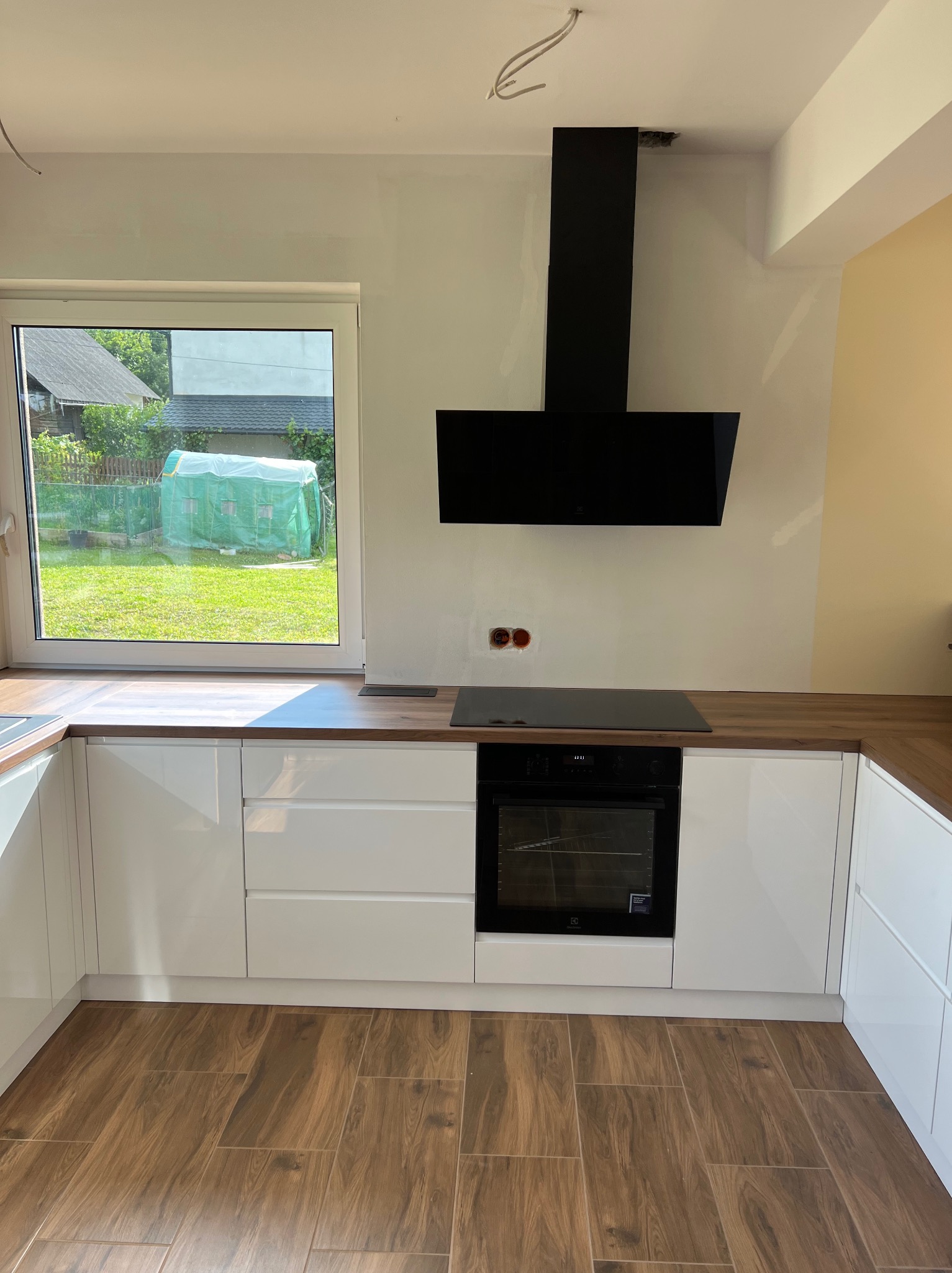 Nowoczesna kuchnia z białymi szafkami, drewnianym blatem i czarnym okapem, widok na ogród przez okno, brązowe płytki podłogowe.