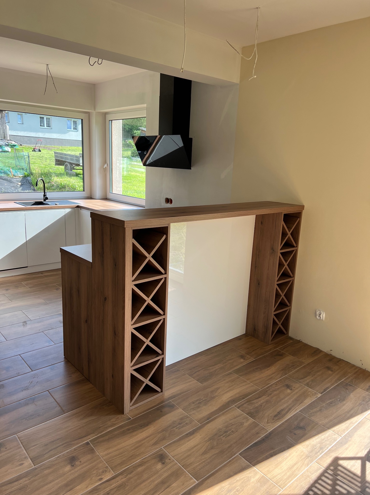 Wnętrze kuchni w trakcie wykańczania z drewnianym blatem, białymi szafkami, czarnym okapem i barkiem z wbudowanym regałem na wino, wykończone płytkami imitującymi drewno na podłodze.