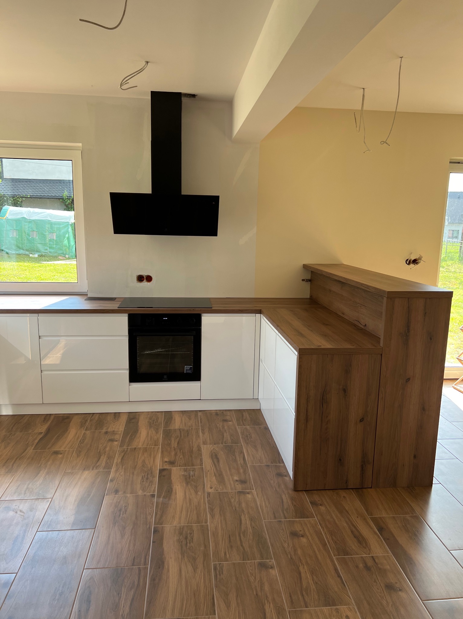 Nowoczesna kuchnia z białymi frontami szafek i drewnianym blatem, czarny okap, płytki podłogowe imitujące drewno, widoczne niedokończone instalacje elektryczne.