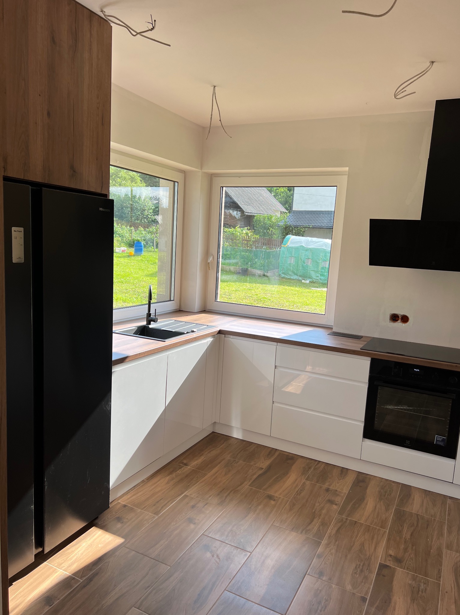 Jasna kuchnia z białymi szafkami, drewnianym blatem i brązową podłogą imitującą drewno, widok na ogród przez okno, widoczne niedokończone prace elektryczne na suficie.