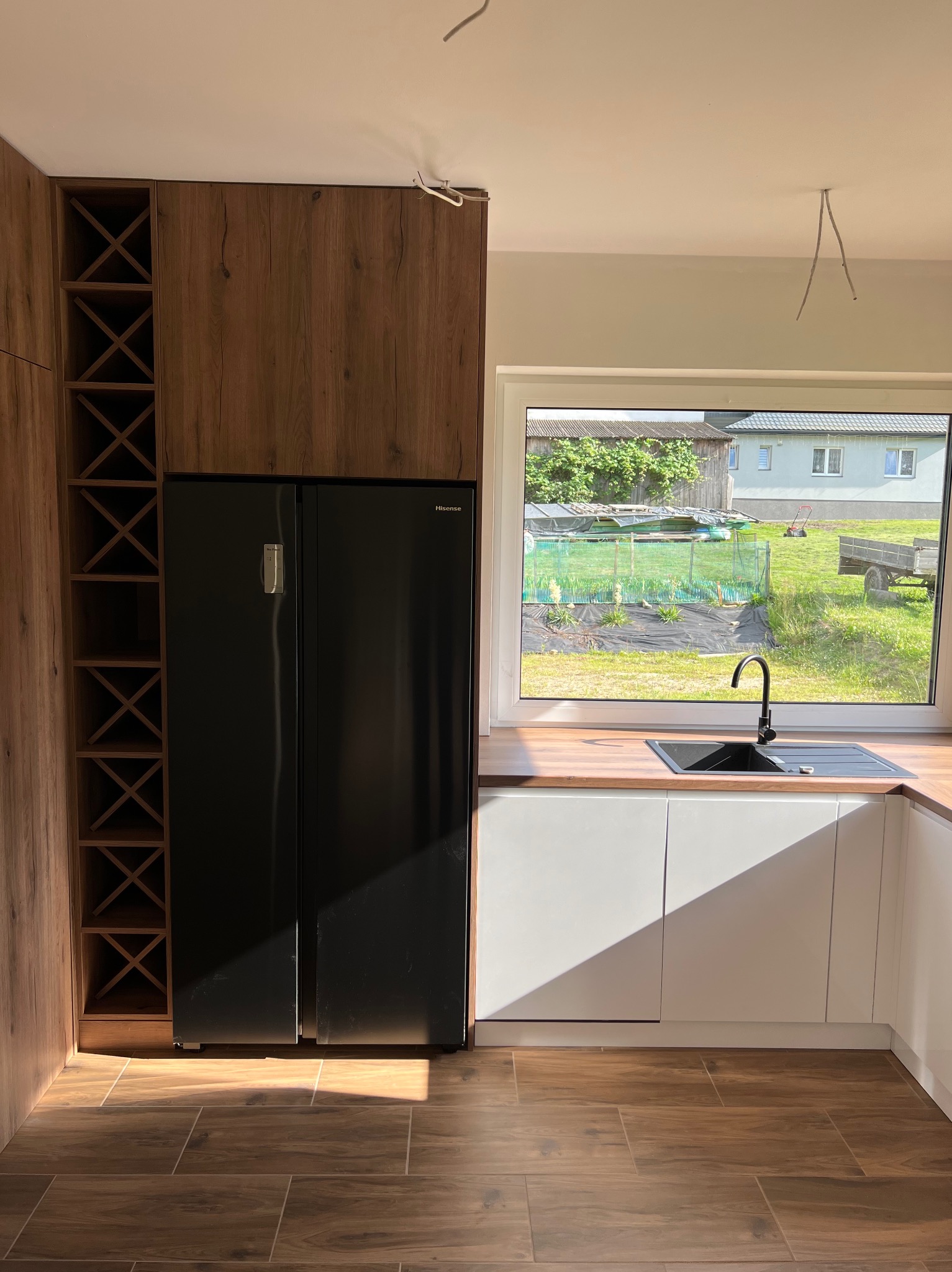 Nowoczesna kuchnia z drewnianymi akcentami: zabudowa z szafkami i półkami na wino w odcieniach brązu, czarna lodówka side-by-side oraz białe szafki dolne z drewnianym blatem i widokiem na ogród...