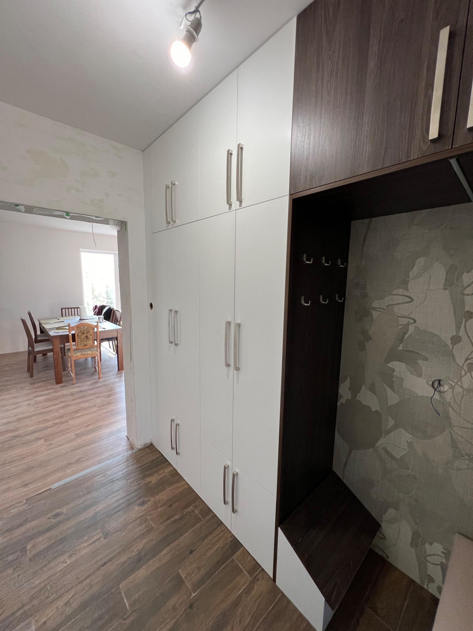 Biało-brązowa zabudowa meblowa z szafkami i otwartą wnęką z wieszakami, widoczna w przedpokoju z drewnopodobną podłogą i przejściem do jadalni w tle.
