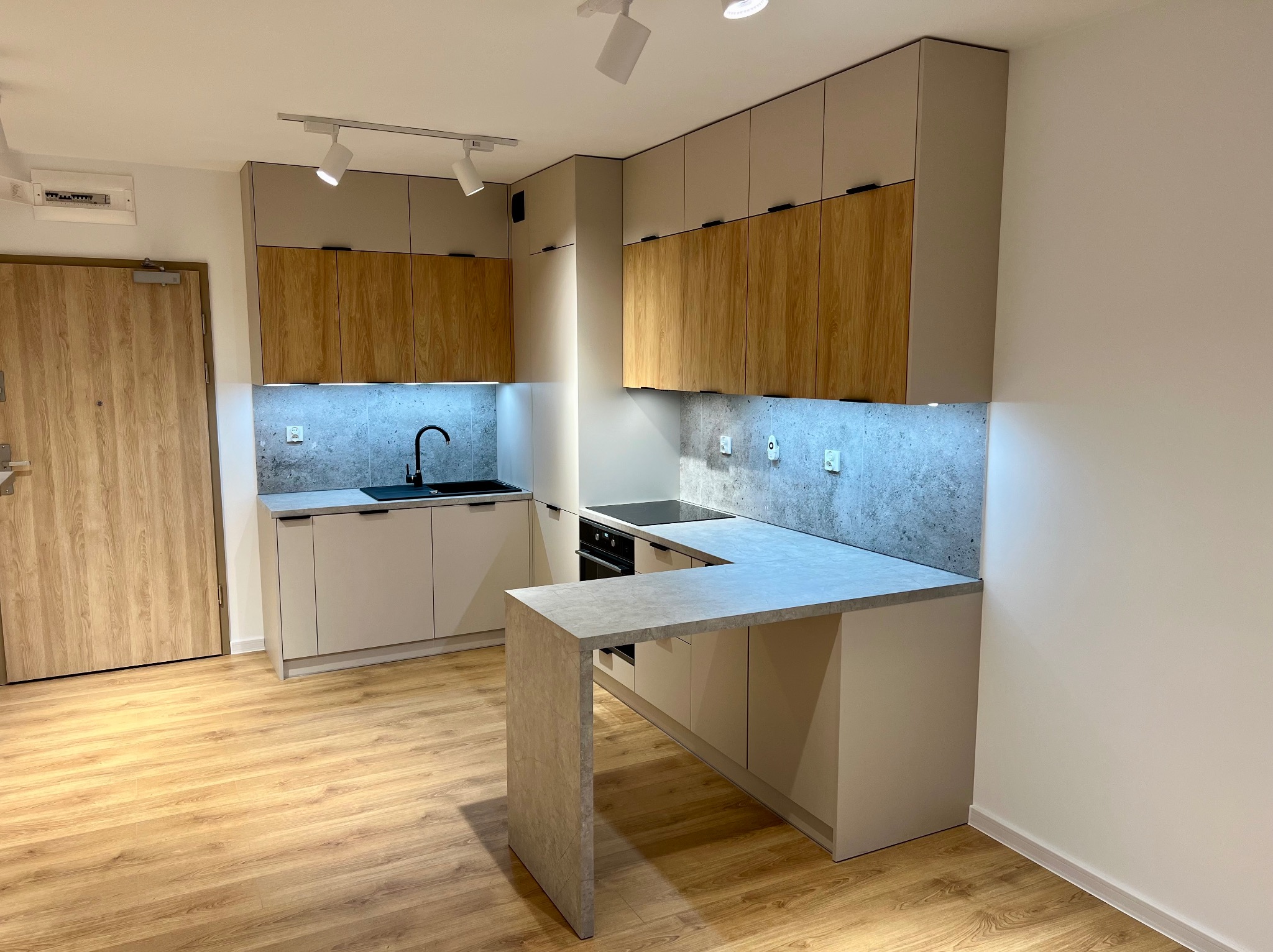 Nowoczesna kuchnia z szarymi frontami, drewnianymi szafkami wiszącymi i blatem imitującym beton, oświetlona punktowymi lampami sufitowymi, z widocznym fragmentem drewnianej podłogi i drzwi.