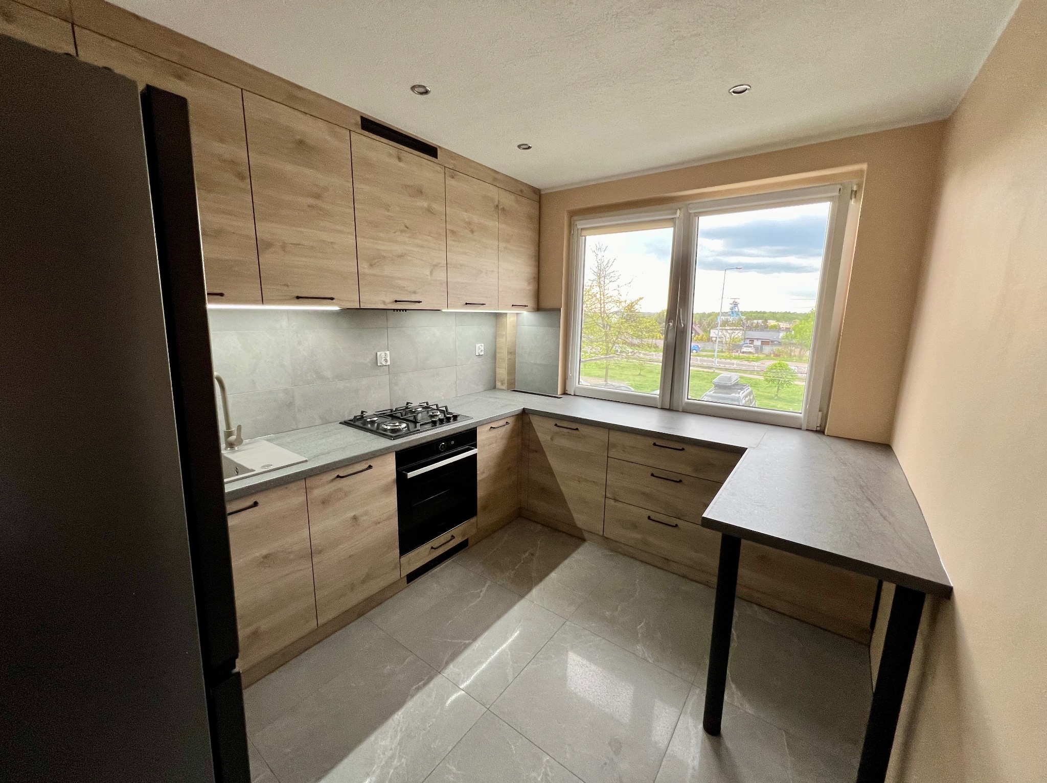 Jasna kuchnia na wymiar z drewnianymi frontami, szarym blatem i płytkami podłogowymi, widok na zewnątrz przez duże okno.