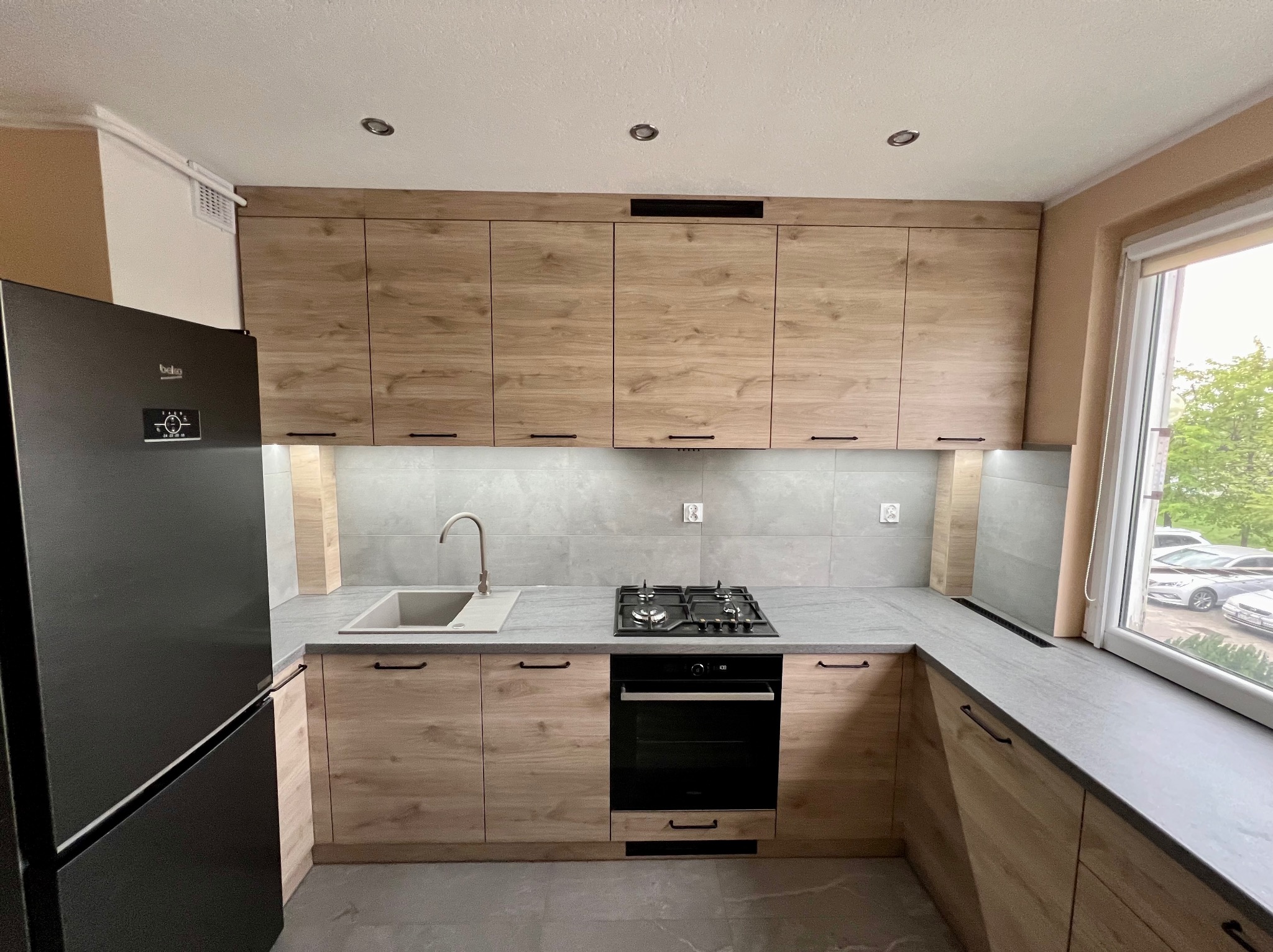 Nowoczesna kuchnia z drewnianymi frontami szafek, szarym blatem i czarnymi uchwytami, widok na okno z widocznymi samochodami na zewnątrz.