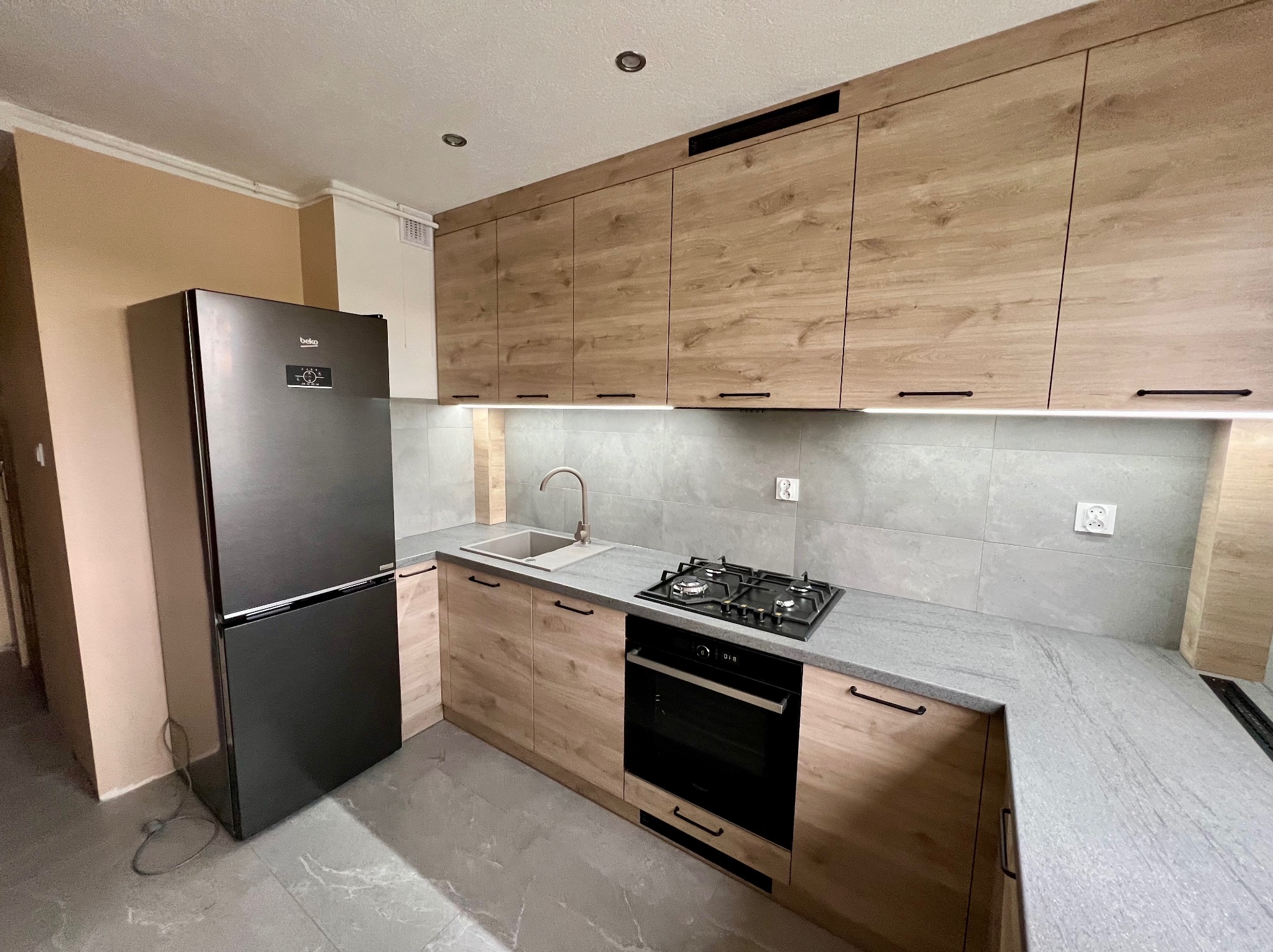 Nowoczesna kuchnia z drewnianymi frontami szafek, szarym blatem i płytkami, czarnym piekarnikiem i lodówką Beko oraz minimalistycznym oświetleniem punktowym.