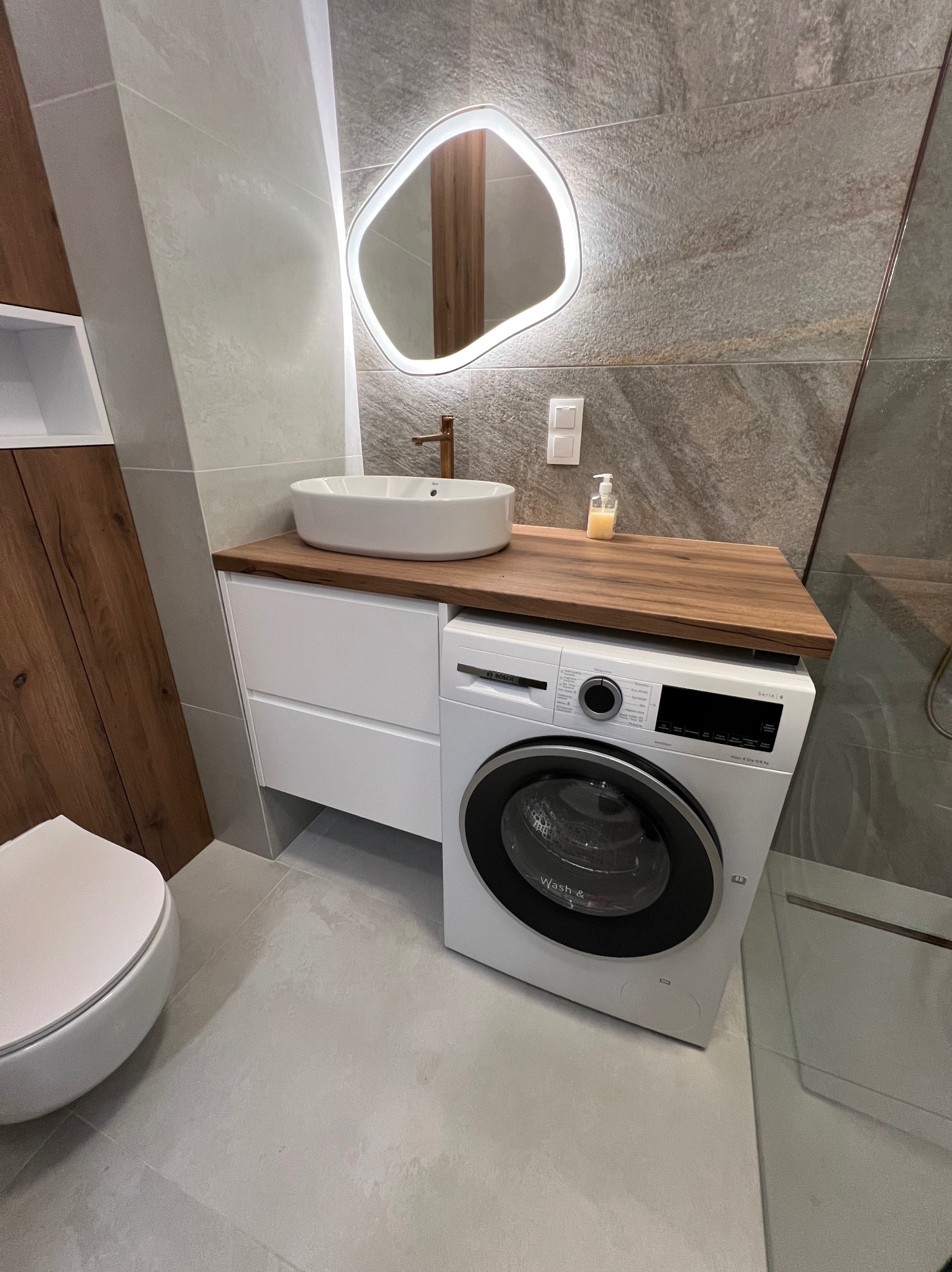 Nowoczesna łazienka z umywalką nablatową na blacie z drewna, pod którym znajduje się pralka; lustro z podświetleniem LED o nieregularnym kształcie.