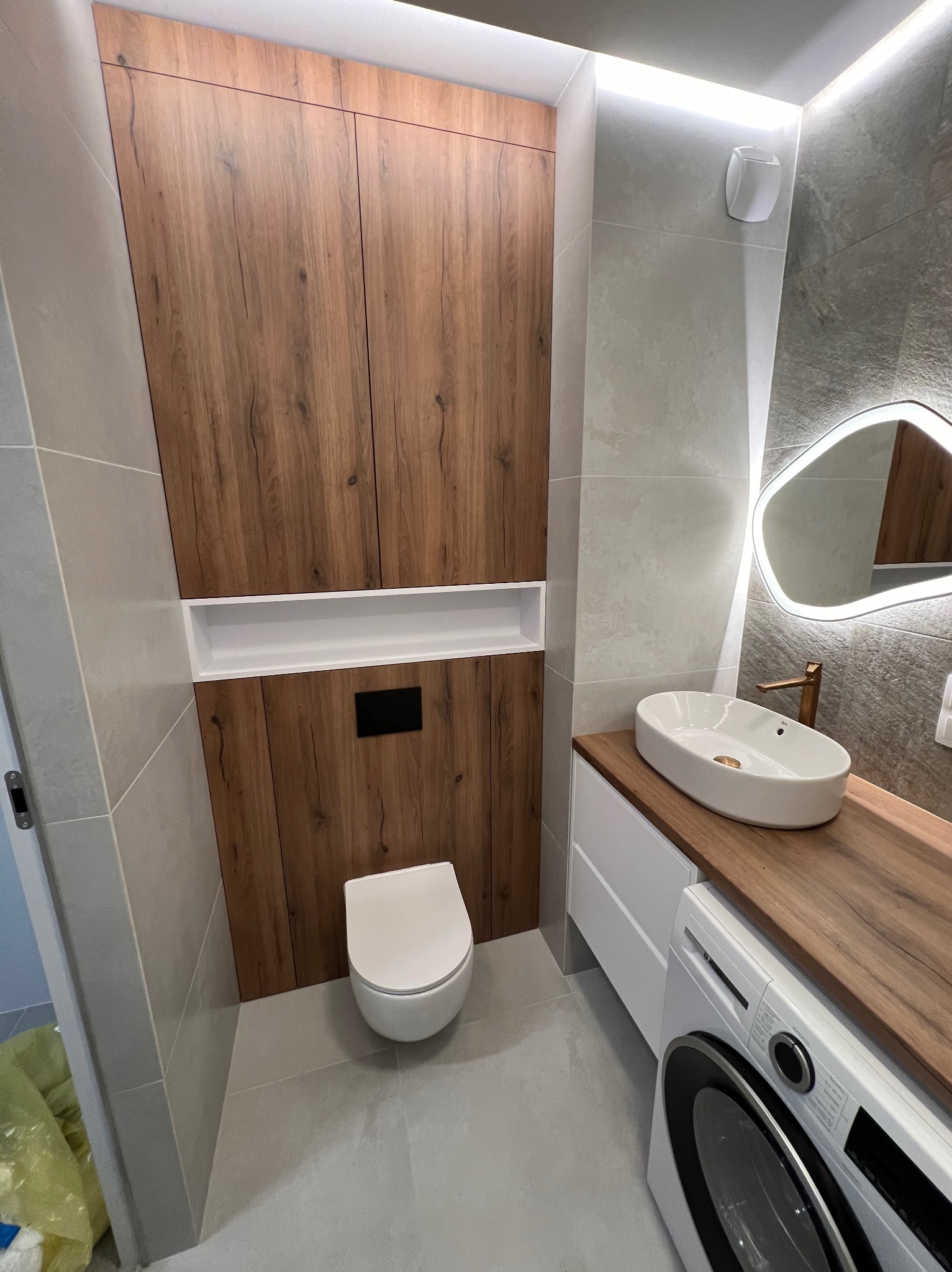 Nowoczesna łazienka z drewnianymi akcentami: szafka wisząca nad toaletą, umywalka nablatowa na blacie z efektem drewna, oświetlone lustro o nieregularnym kształcie i pralka pod blatem.