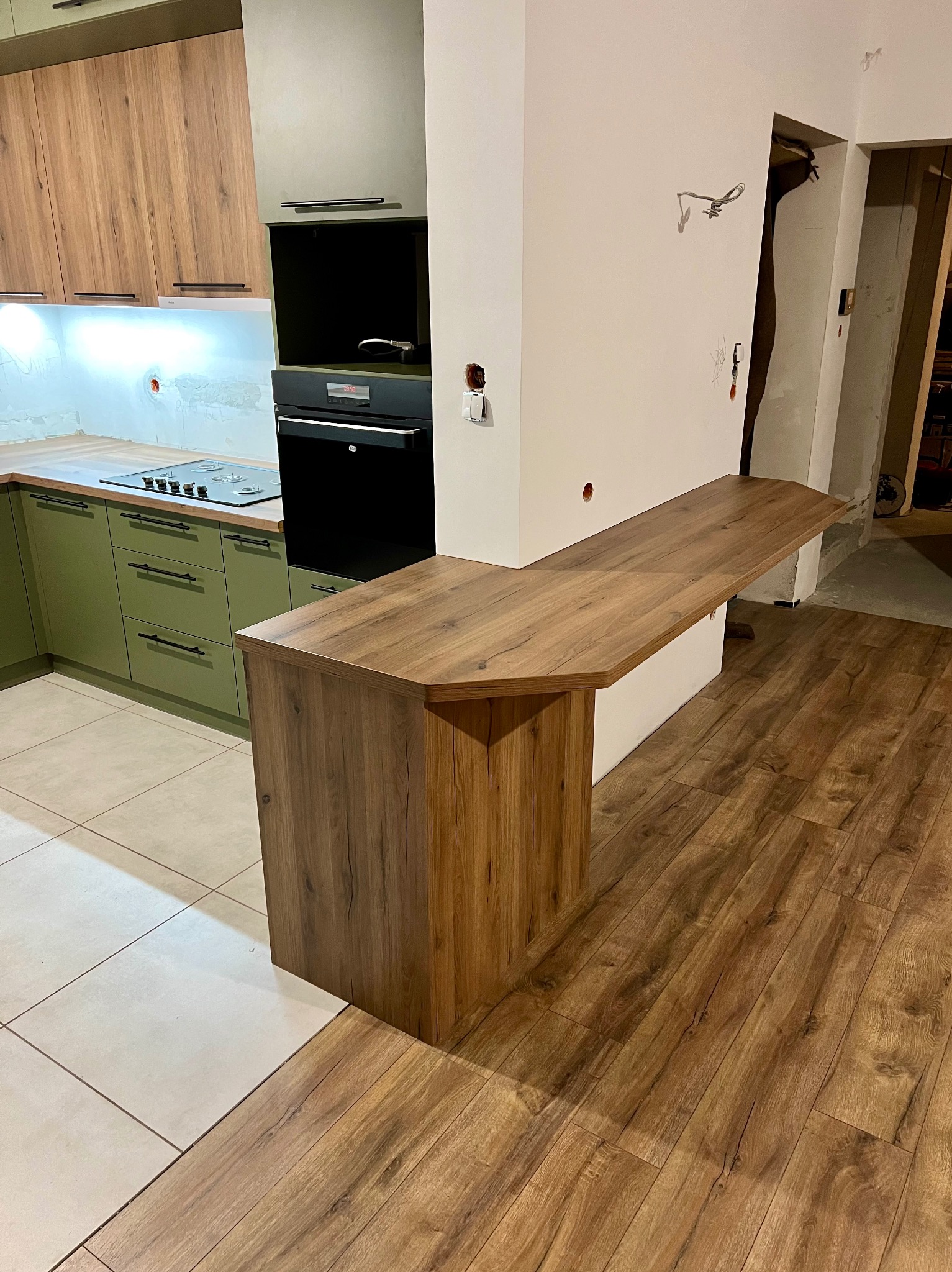 Nowoczesna kuchnia z zielonymi szafkami i drewnianym blatem w Budzowie, z widocznym barkiem oddzielającym przestrzeń kuchenną od salonu, podłoga wyłożona panelami.